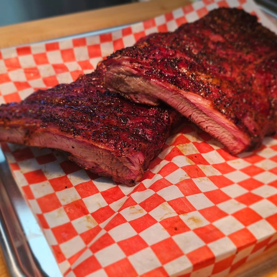 Two racks of Arctic Smokehouse BBQ ribs with a dark, seasoned crust on red and white checkered paper.