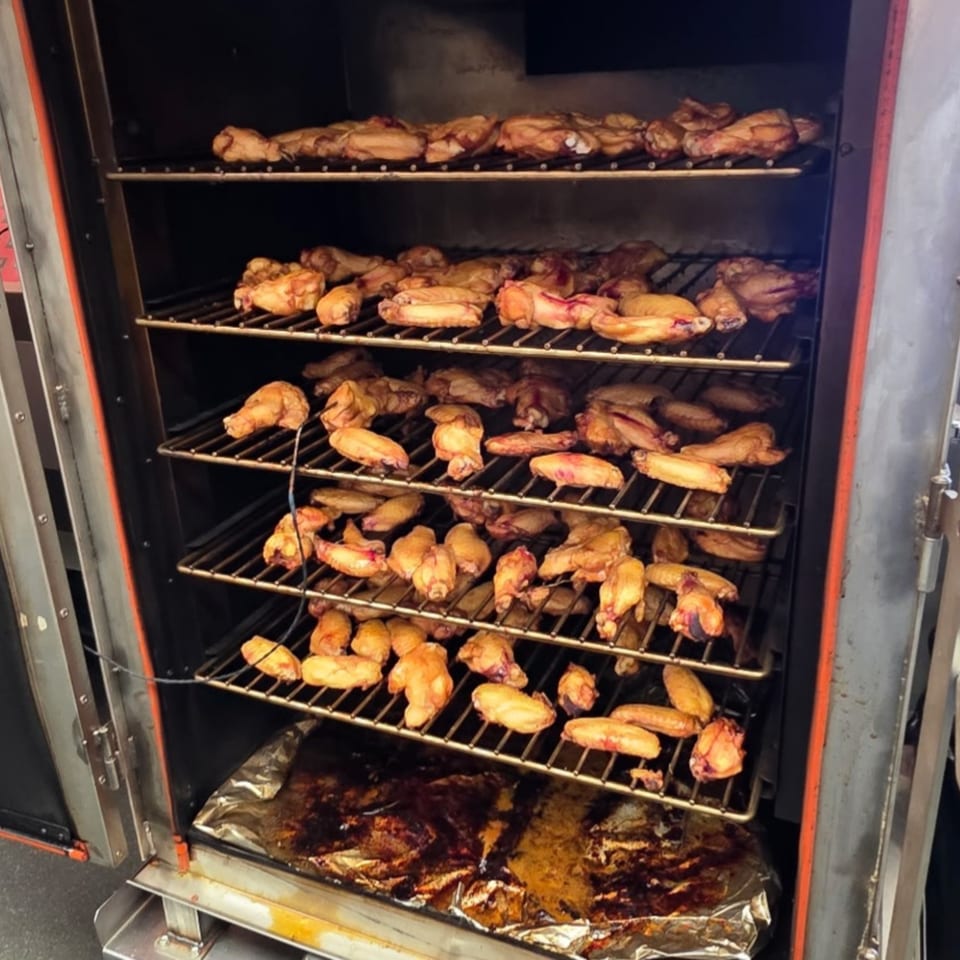 Chicken wings cooking on multiple racks inside a large, dark smoker at Arctic Smokehouse BBQ.