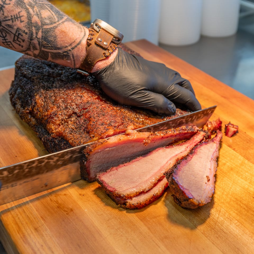 A gloved hand slices a large, smoked brisket on a wooden cutting board, revealing juicy pink meat.